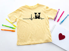 Load image into Gallery viewer, Yellow t-shirt with a design of two cats holding a heart, placed on a white surface with colorful pens and a red heart.