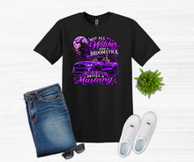Load image into Gallery viewer, A black graphic t-shirt displayed in a flat-lay arrangement with folded blue jeans, white sneakers, sunglasses, and a small green plant on a light wood background. The t-shirt features a purple graphic of a witch riding a purple Mustang with the text 'NOT ALL Witches RIDE A BROOMSTICK ONE OF THE DRIVES A Mustang'.