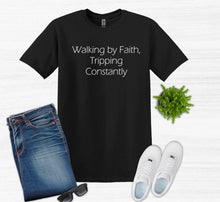 Load image into Gallery viewer, Black t-shirt with text 'Walking by Faith, Tripping Constantly', paired with jeans, sneakers, and sunglasses on a white background.