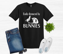Load image into Gallery viewer, Black t-shirt with 'Easily distracted by bunnies' text and graphics, paired with jeans, sneakers, and sunglasses on a white background.