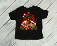 Load image into Gallery viewer, A black short-sleeve t-shirt laid flat on a light gray wood plank background. The front of the shirt features a colorful graphic of two guinea pigs wearing Santa hats under a Christmas tree with the text 'Wheeking around the Christmas Tree'.