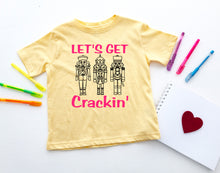 Load image into Gallery viewer, A pale yellow short-sleeve T-shirt laid flat on a white surface, printed with the phrase 'LET'S GET Crackin'' in bright pink and three black nutcracker figures. Colorful pens, a white notepad with a red heart, and a small decorative object are placed around the shirt as styled props.