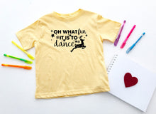 Load image into Gallery viewer, A pale yellow short-sleeve t-shirt with a black graphic text reading 'OH WHAT fun IT IS TO dance' and a dancing reindeer, laid flat on a white background with colorful pens, a notepad, and a red heart.