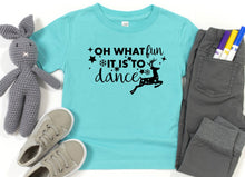 Load image into Gallery viewer, A flat-lay of a turquoise short-sleeve t-shirt with a black graphic that reads 'OH WHAT fun IT IS TO dance' with stars, snowflakes, and a deer silhouette. The shirt is styled with gray pants, gray sneakers, a gray crocheted bunny toy, and markers on a white background.