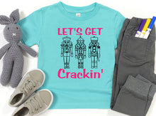 Load image into Gallery viewer, A turquoise short-sleeve T-shirt laid flat with a pink and black graphic reading 'LET'S GET Crackin'' and three nutcracker illustrations. The shirt is styled in a flat lay with gray pants, a gray crocheted bunny toy, beige sneakers, and colored markers on a white background.