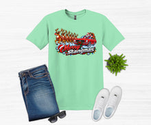 Load image into Gallery viewer, A mint green graphic t-shirt featuring a 'Merry Stangmas' Santa Claus in a red race car with reindeer design, styled in a flat lay with blue jeans, white sneakers, sunglasses, and a small green plant on a white wood background.