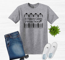 Load image into Gallery viewer, A heather gray short-sleeve T-shirt with a black graphic reading 'St. Frances Cabrini Preschool' and schoolhouse illustrations, styled in a flat lay with blue jeans, white sneakers, sunglasses, and a small green plant on a light background.