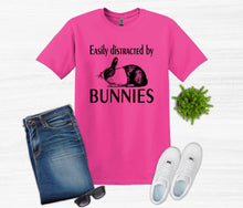 Load image into Gallery viewer, Pink t-shirt with 'Easily distracted by bunnies' text and graphics, paired with jeans, sneakers, and sunglasses on a light gray background.