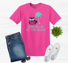 Load image into Gallery viewer, Pink t-shirt with 'Chinchillin' at the Beach' design, blue jeans, white sneakers, and sunglasses on a white background.