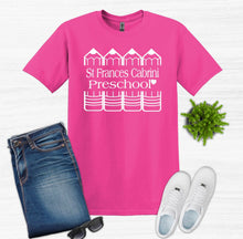 Load image into Gallery viewer, A bright pink short-sleeve T-shirt with a white printed graphic reading 'St. Frances Cabrini Preschool' and small house/crib illustrations, shown in a flat-lay with blue jeans, white sneakers, sunglasses, and a small green plant on a light wood background.