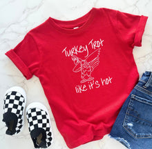 Load image into Gallery viewer, A red short-sleeve t-shirt with a white graphic of a turkey and the text 'Turkey Trot like it's hot', styled in a flat lay with black and white checkered slip-on shoes and blue jeans on a light marble background.