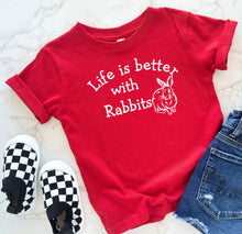 Load image into Gallery viewer, A red short-sleeve graphic T-shirt laid flat on a light marble surface, with white text reading 'Life is better with Rabbits' and a white rabbit illustration. Black-and-white checkered slip-on shoes and blue denim shorts are styled around it.