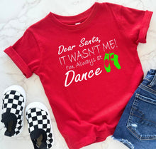 Load image into Gallery viewer, A red short-sleeve graphic T-shirt laid flat on a light marble surface, styled with black-and-white checkered slip-on shoes and blue jeans. The shirt reads, 'Dear Santa, IT WASN'T ME! I'm Always at Dance' in white script and block lettering, with bright green ballet slippers graphic.