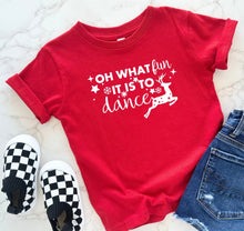 Load image into Gallery viewer, A red short-sleeve graphic T-shirt laid flat on a light marble surface, styled with black-and-white checkered slip-on shoes and blue denim jeans. The shirt has white text reading “OH WHAT fun IT IS TO dance” with small star and snowflake motifs and a white reindeer graphic.