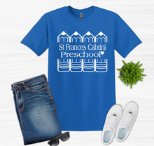 Load image into Gallery viewer, A royal blue short-sleeve t-shirt with a white graphic reading 'St Frances Cabrini Preschool' and schoolhouse illustrations, styled in a flat lay with folded blue jeans, white sneakers, sunglasses, and a small green plant on a white wood-textured background.