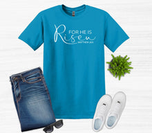 Load image into Gallery viewer, Blue t-shirt with 'For He is Risen' text, jeans, white sneakers, sunglasses, and a plant on a light wooden surface.