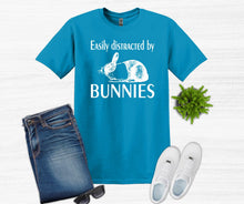 Load image into Gallery viewer, Blue t-shirt with 'Easily distracted by bunnies' text and graphics, paired with jeans, sunglasses, and shoes on a white background.