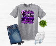 Load image into Gallery viewer, A heather gray graphic t-shirt featuring a purple design with a mustang car and the text 'Not All Witches Ride Broomsticks One of them Drives a Mustang', styled in a flat lay with blue jeans, white sneakers, sunglasses, and a small green plant on a white wood background.