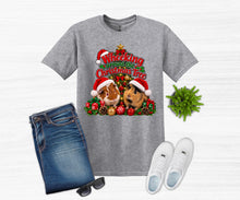 Load image into Gallery viewer, A heather gray graphic t-shirt featuring two guinea pigs wearing Santa hats and the text 'Wheeking Around the Christmas Tree', styled in a flat lay with blue jeans, white sneakers, sunglasses, and a small green plant on a white wood-textured background.