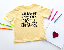 Load image into Gallery viewer, A pale yellow short-sleeve T-shirt laid flat on a white background, printed with the phrase 'We'll you a Merry Christmas' in black lettering with paw-print and star accents. Colorful pens, a white notepad with a red heart, and other small stationery items are arranged around it.