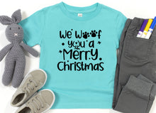 Load image into Gallery viewer, A turquoise short-sleeve T-shirt laid flat with black text reading 'We Woof You a Merry Christmas' and paw-print/star accents. The shirt is styled with a gray stuffed bunny, gray pants, and gray sneakers on a white background.