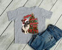Load image into Gallery viewer, A heather gray short-sleeve t-shirt with a Christmas graphic of a rabbit in a Santa hat next to a decorated Christmas tree and the text 'Hopping Around the Christmas Tree', styled flat-lay with blue jeans on a white wood plank background.