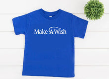 Load image into Gallery viewer, A royal blue short-sleeve t-shirt with 'Make A Wish' printed in white across the chest, displayed flat on a white wood plank background with a small green plant in the top right corner.