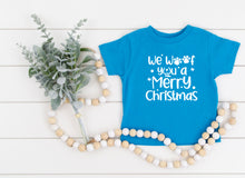 Load image into Gallery viewer, A bright blue short-sleeve t-shirt with white text reading 'We Woof you a Merry Christmas' and paw prints, styled in a flat lay with a green eucalyptus sprig and a wooden bead garland on a white plank background.