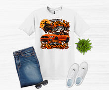 Load image into Gallery viewer, A white graphic T-shirt laid flat on a light wood background, styled with folded blue jeans, black sunglasses, white sneakers, and a small green plant. The shirt features a large autumn-themed graphic with the text 'Not All Witches Ride a Broomstick One of them Drives a Mustang' around an orange convertible car and a witch silhouette.