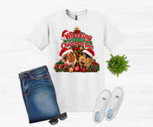 Load image into Gallery viewer, A white graphic t-shirt featuring a festive Christmas tree design with two guinea pigs in Santa hats, styled in a flat lay with blue jeans, white sneakers, sunglasses, and a small green plant on a white wood-textured background.